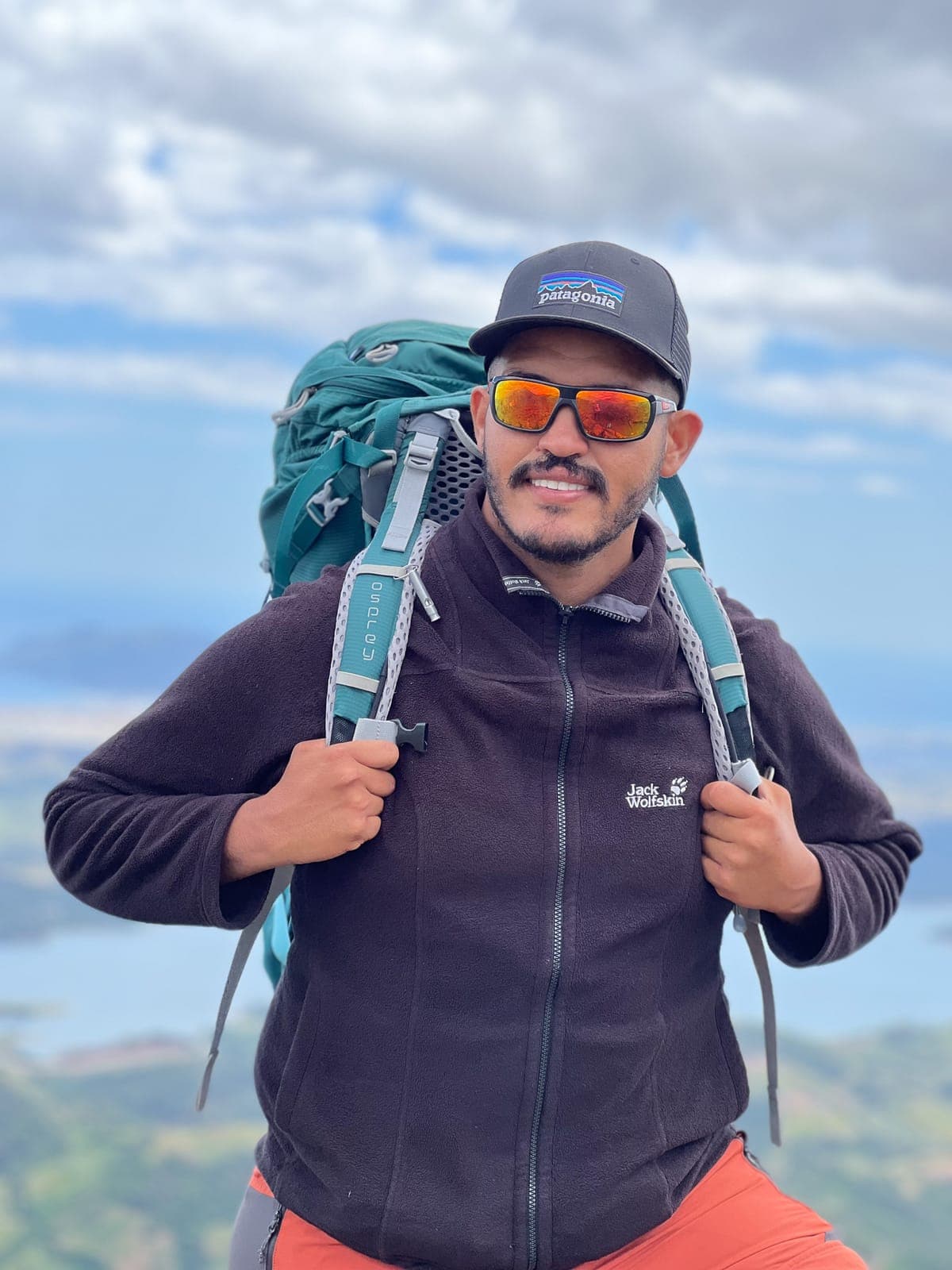 Mounir on a mountain top, wearing a trekking backpack, with blue water and green hills far below
